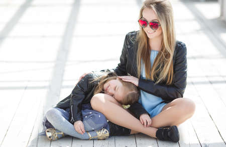 Beautiful woman in red mirrored glasses in the shape of hearts, blonde with long blond hair in a black leather jacket, spending time outdoors in the spring with his blond daughter, a girl of four years old, wearing a black leather jacketの写真素材