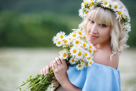 Summer portrait of a happy woman on a white field of blooming daisies in a mountainous area, a woman, a blonde with a beautiful hairstyle and light make-up, ears are silver earrings, dressed in a sundress of blue in a wreath of white daisiesの写真素材