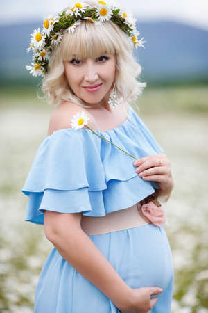 Spring portrait of a pregnant woman on a white field of blooming daisies in a mountainous area, a woman with long blonde eyelashes and light make-up, ears are silver earrings, large belly, dressed in a sundress of blue in a wreath of white daisiesの写真素材