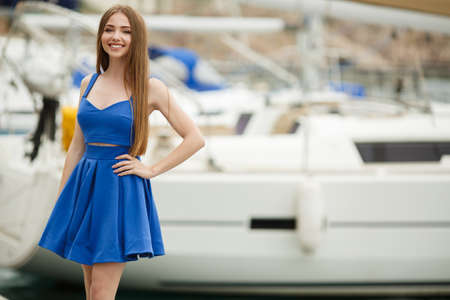 Slender blond young woman with gray-green eyes and long straight hair, dressed in a blue sundress, a beautiful smile and light makeup posing outdoors in the summer against white yachts and boats on the dockの写真素材