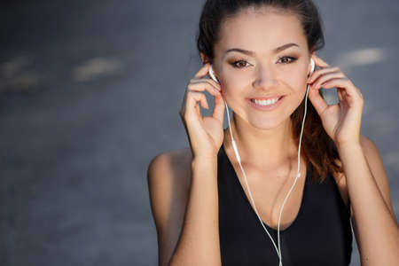 Portrait of a beautiful young woman, brunette with brown eyes, a beautiful smile and straight white teeth, wearing a black T-shirt, listening to music on white earphones outdoors in summer parkの写真素材