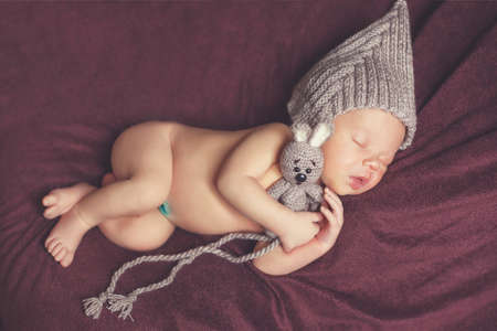 Closeup portrait of newborn baby in gray knitted hat with a grey toy Bunny, a little fluffy hair, sweetly asleep on a dark brown blanket, lying on tummy, put hands behind his head and legs under herの写真素材