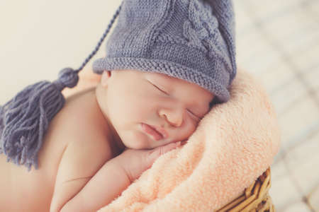 Adorable little guy with little fluffy hairs, in a gray knitted cap, tucked under his legs and put the handle under his head, sound asleep on the soft beige blanket in a large wicker basket high, sweet sleeping newborn babyの写真素材