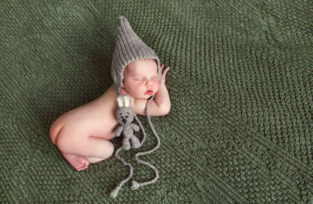 Closeup portrait of newborn baby in gray knitted cap with grey a toy mouse, little furry hairs, sweetly asleep on a dark green blanket lying on tummy, put hands below the head and legs tucked under herselfの写真素材