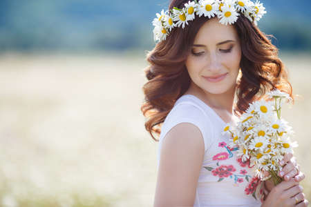 Cute pregnant brunette woman in white t-shirt with a bouquet of white daisies on his head wears a wreath of white flowers, chamomile, summer, walking alone in a beautiful field of blooming white daisiesの写真素材