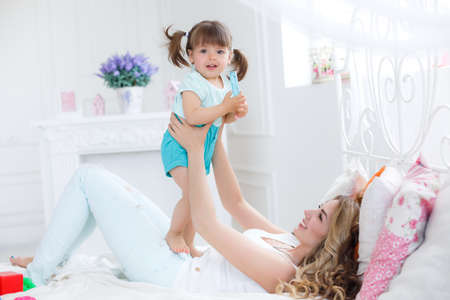 Happy young mother, blonde with long curly hair playing with his little daughter, a brunette, spend time in bright bedroom on a white bed, both dressed in white panties and white tops, having fun and smilingの写真素材