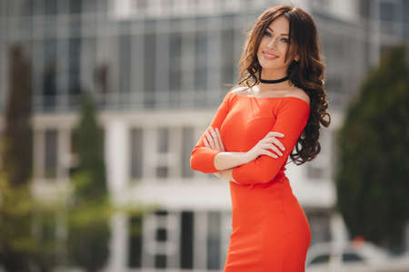 Summer portrait of a beautiful slim woman, brunette with long curly hair and grey eyes, dressed in a bright orange dress, wearing large black earrings, a cute smile posing outdoors in the city in the summerの写真素材