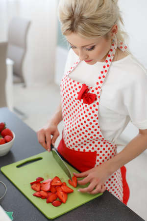 Young beautiful woman, blonde hair, light makeup and pink lipstick, wearing earrings, wearing a white t-shirt and white red polka dot pinafore, engaged in the bright kitchen cutting red strawberries to decorate cakesの写真素材