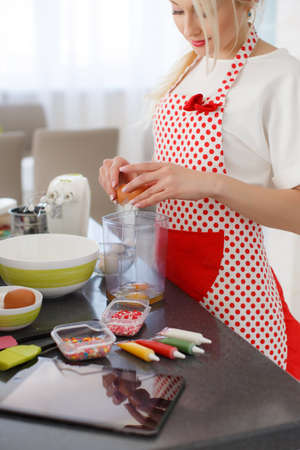 Young beautiful woman, blonde hair, light makeup and pink lipstick, nice smile, wears earrings, wears a white t-shirt and white red polka dot pinafore, engaged in the bright kitchen cooking bakingの写真素材