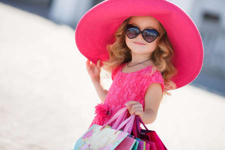 Beautiful little girl with blond long curly hair, in a pink straw hat with large brim, sun glasses with glass in the form of hearts, in a pink dress with a bunch of colored shopping bags, posing near a supermarketの写真素材