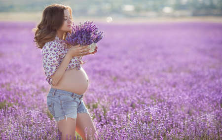 Beautiful pregnant brunette woman with long curly hair, big belly, wearing a light shirt and blue shorts, a hand holding a basket of flowers, picking flowers on summer mountain meadow of lavender in summerの写真素材