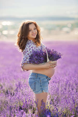 Beautiful pregnant brunette woman with long curly hair, big belly, wearing a light shirt and blue shorts, a hand holding a basket of flowers, picking flowers on summer mountain meadow of lavender in summerの写真素材