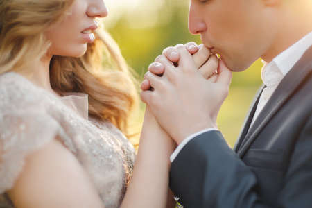 Young loving couple on the wedding day, the groom-a man with a short haircut, in a dark wedding suit and a beautiful bride-a woman with long curly hair in beige wedding dress standing arm in arm at sunset in summer green Parkの写真素材