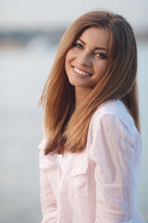 Portrait of a beautiful woman, brunette with long straight hair and gray eyes, a nice smile and beautiful white teeth, dressed in a white shirt resting on the shore of the blue ocean in the summer in the fresh airの写真素材