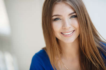 Beautiful young woman brunette with long straight hair, long black eyelashes, pretty smile, white straight teeth, wearing a blue summer dress, wears a gold chain on the neck, posing on light gray backgroundの写真素材