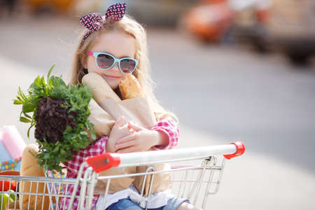 The little blonde girl with black and pink bow on head, wearing sun glasses with a blue frame in a red plaid shirt, sitting in a cart filled with groceries at the supermarket, clutching a paper bag with the loaves and greensの写真素材