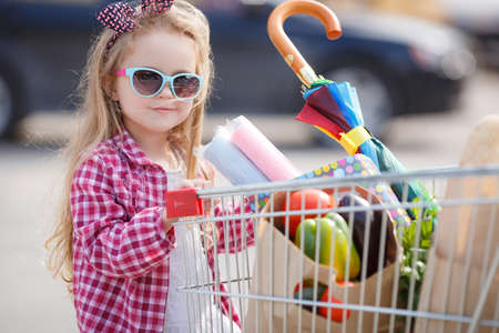 The little blonde girl with bow on her head, wears sunglasses in blue frame, dressed in a plaid shirt is a red and white, standing near the basket on wheels, loaded with food and shoppingの写真素材