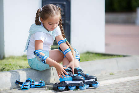 Blonde little girl with two long pigtails wearing a white t-shirt and blue shorts, wearing knee pads and protection on elbows blue, spends time alone in a city Park, roller skating black and blue in the summerの写真素材