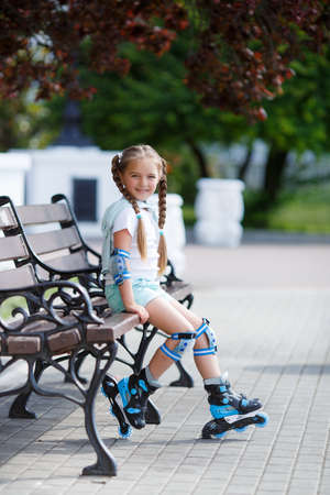 Blonde little girl with two long pigtails wearing a white t-shirt and blue shorts, wearing knee pads and protection on elbows blue, spends time alone in a city Park, roller skating black and blue in the summerの写真素材