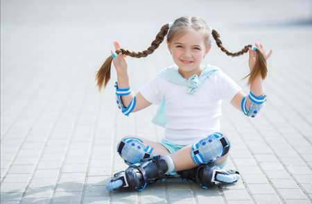 Blonde little girl with two long pigtails wearing a white t-shirt and blue shorts, wearing knee pads and protection on elbows blue, spends time alone in a city Park, roller skating black and blue in the summerの写真素材