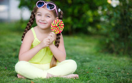 Little girl, brunette with long hair braided in two pigtails, in the ears of gold earrings, wearing sun glasses in a pink frame with glass in the shape of hearts, wearing a yellow T-shirt and pants, sitting on green grass in the summer park with a large rの写真素材