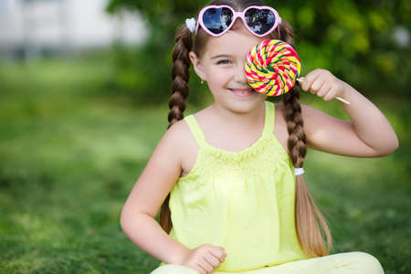 Little girl, brunette with long hair braided in two pigtails, in the ears of gold earrings, wearing sun glasses in a pink frame with glass in the shape of hearts, wearing a yellow T-shirt and pants, sitting on green grass in the summer park with a large rの写真素材