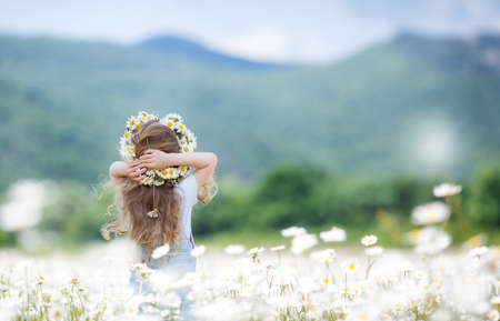 Cute little girl with curly, long hair, dressed in light blue overalls and a blue shirt with white polka dots, on the head wears a wreath of white wildflowers posing on a blooming white daisies field in a mountainous area in the summer, rear viewの写真素材