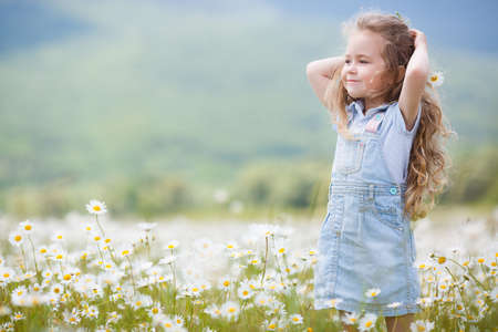 Cute little girl with brown eyes and curly long hair, dressed in light blue overalls and a blue shirt with white polka dots, posing on a blooming white daisies field in a mountainous area in the summerの写真素材