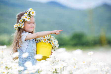 Beautiful little girl with long curly blond hair, cute smile, in a light blue denim overalls, a white wreath of fresh flowers in the hands holding a yellow bucket with a field of daisies, picking flowers on a mountain meadow in summerの写真素材