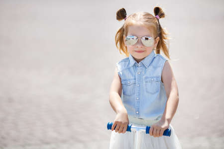 Little girl with blond hair, hair in two ponytails, in mirrored sun glasses, a light blue sleeveless shirt and white skirt, the one riding on the scooter in the city in the fresh air in summerの写真素材