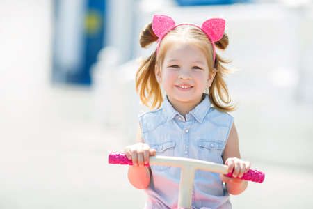 Little girl with blond hair, hair in two tails, color is pink hoop hair with pink ears, wearing a light blue sleeveless shirt and pink skirt, riding a pink bicycle in the children in the city of the great outdoors in the summerの写真素材