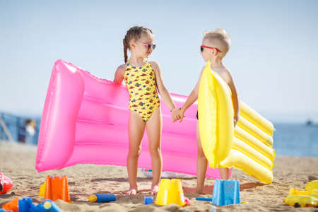 Little girl with pigtails and a little blond boy with short hair, wearing dark sun glasses, spend time together on the sandy beach, standing close, holding a yellow and pink mattress for swimming, posing against the backdrop of the oceanの写真素材