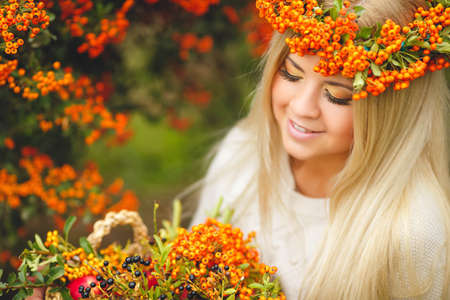 Young beautiful woman with long blonde straight hair, light makeup, wearing a beige jacket, on the head wears a wreath of orange of Rowan berries, spending time outdoors in the Park in the middle of autumn, posing near bushes ripe Rowanの写真素材