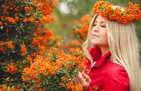 Young beautiful woman with long blonde straight hair, light makeup, wearing a red jacket, on the head wears a wreath of orange of Rowan berries, spending time outdoors in the Park in the middle of autumn, posing near bushes ripe Rowanの写真素材