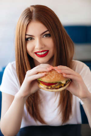Beautiful woman with red long straight hair, bright make-up, brown eyes, red lipstick, long eyelashes, pink nail polish in the hands holding a large hamburger, sitting at a table in a cafe, dinner aloneの写真素材