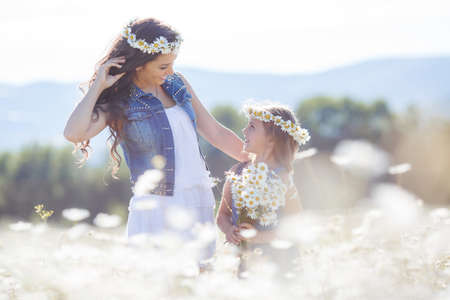 Cheerful family of two people, mother and little daughter spend time together in the highlands on a field of blooming white daisies in summer in the fresh air, mother and daughter wearing wreaths of flowers-white daisies, both brunettes, girl with pigtailの写真素材