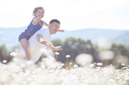 Cheerful family of two persons, father and little daughter spend time together in the highlands on a field of blooming white daisies in summer in the fresh air, a girl dressed in blue overalls and wears a wreath of wild flowers-white daisiesの写真素材