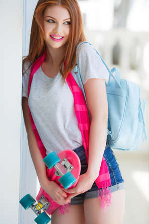 Beautiful young woman with long straight red hair, light makeup, brown eyes, lipstick pink color, wearing a light gray t-shirt and blue denim shorts on the shoulders draped a pink shawl, holding a pink skateboard, posing in the summer outdoors in the cityの写真素材