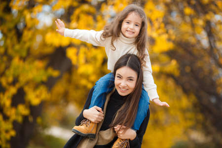 A beautiful young woman of Eastern appearance, brown hair, brown eyes, pleasant smile, dressed in a black jacket and blue jeans, holding on the shoulders of his little daughter, a girl with blond hair and brown eyes, walking together in yellow autumn Parkの写真素材