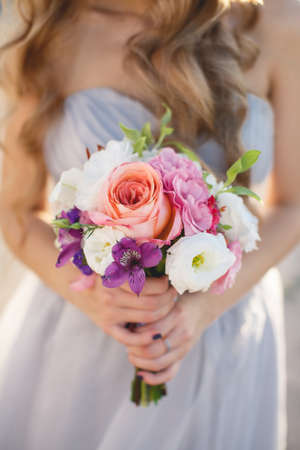 Beautiful wedding bouquet of assorted colors of white, pink, lilac and red colors in the hands of the bride dressed in white wedding dress, curly blond hair, the attention is focused on the bouquet of flowersの写真素材