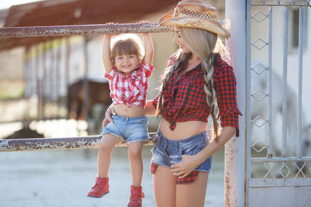 Beautiful young woman with long blonde hair braided in two braids, a cowboy hat, spends time in the countryside in the fresh air in summer with his young daughter, the girl blonde, mom and daughter dressed in red checkered shirt and blue denim shortsの写真素材