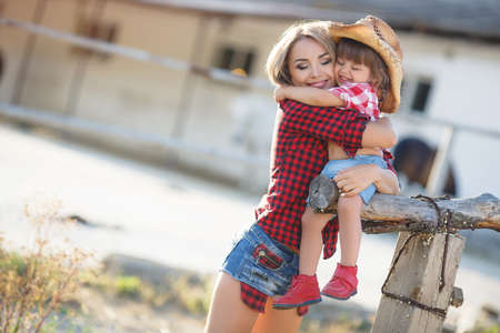 Happy young mother, a woman with long blonde straight hair, spends time in the village in the summer with his daughter, a little girl with blond hair and mother and daughter are dressed in red checkered shirt and blue denim shortsの写真素材