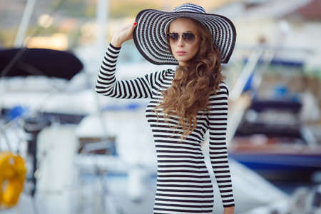 Beautiful young woman model looks, brunette with long curly hair, wears dark sun glasses on his head wearing striped straw hat with a large brim, dressed in a striped summer dress, spends time on the ocean, close to Parking of boats and yachtsの写真素材