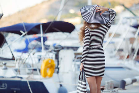 Beautiful young woman model looks, brunette with long curly hair, on his head wearing striped straw hat with a large brim, dressed in a striped summer dress, spends time on the ocean, close to Parking boats and yacht, rear viewの写真素材