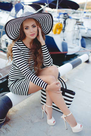 Beautiful young woman model looks, brunette with long curly hair and blue eyes, on his head wearing striped straw hat with a large brim, dressed in a striped summer dress, spends time on the ocean, close to Parking of boats and yachtsの写真素材
