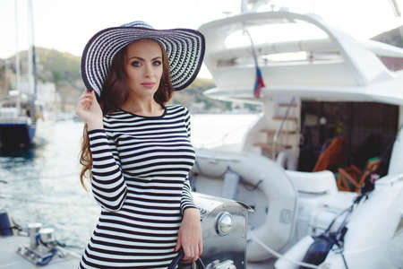 Beautiful young woman model looks, brunette with long curly hair and blue eyes, on his head wearing striped straw hat with a large brim, dressed in a striped summer dress, spends time on the ocean, close to Parking of boats and yachtsの写真素材
