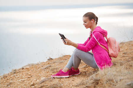 Beautiful brunette woman with long hair braided in a braid, dressed in gray sweat pants, pink sneakers and pink sports jacket with light pink backpack, sitting on a rock on a background of blue sea, communicating in social networks using the tabletの写真素材
