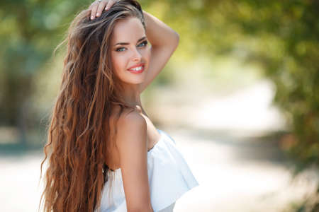 Summer portrait, of beautiful woman with grey eyes, long wavy hair, brown hair, light makeup and pink lipstick, nice smile and white teeth, posing outdoors on the walk to the green Park in Sunny dayの写真素材