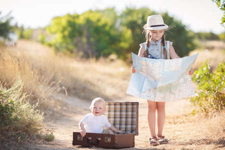 Little boy in a white t-shirt is resting sitting in a large open brown suitcase on a country road, the elder sister is studying map of the area, a girl 6 years old in a gray summer suit with long braids, in a light hat and a beige backpackの写真素材