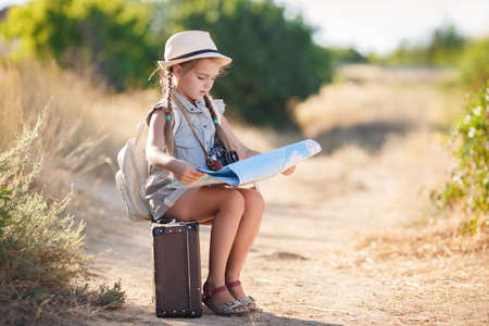 Cute little girl 6 years old, long hair braided into two pigtails, dressed in a summer Onesie blue color, on his head wearing a sun hat, behind wears light leather backpack, sitting on the old suitcase among the back roads, exploring the map of the areaの写真素材